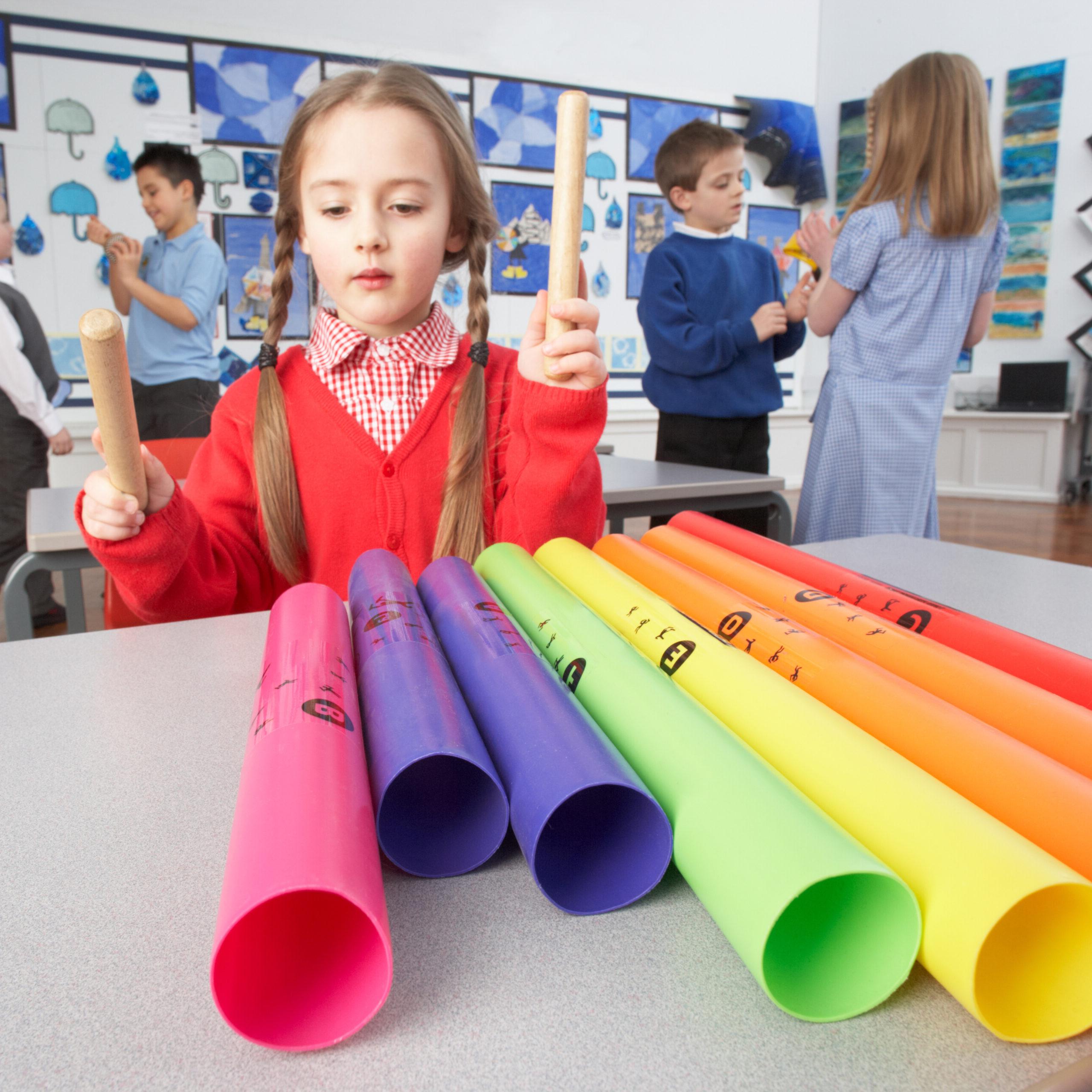 A young girl in a red sweater plays Boomwhackers in a classroom, tapping rhythm sticks on colorful plastic tubes in front of her.