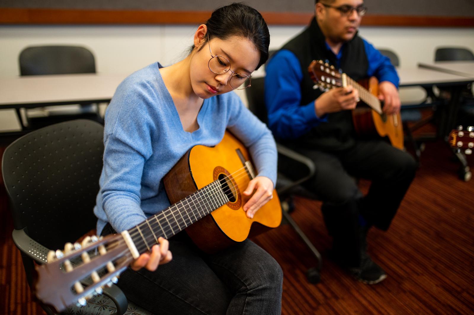 A female and male students, playing the guitar in a classroom setting.